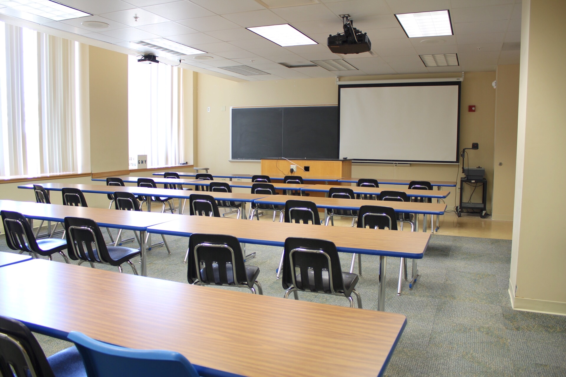 Hoyt讲座104 from back to front of the room shows tables, chairs, blackboard, av cart, ceiling mounted projector, overhead projector on a cart and a projection screen.