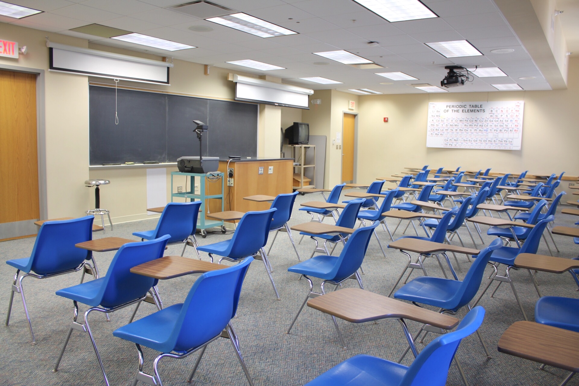Hoyt讲座105 view from the back of the classroom showing chairs with desk arms, 2 ceiling mounted projection screens, ceiling mounted projector, overhead projector on a cart, stationary cabinet, tv on a cart, 2 exit doors, large blackboard.